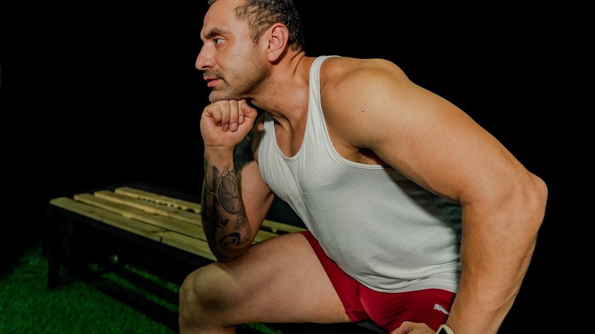 Muscular man in white tank top and red shorts poses thoughtfully on gym bench.